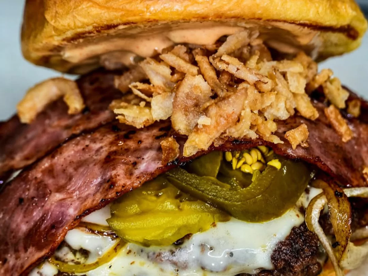 A detailed, close-up shot of a juicy gourmet burger, featuring a beef patty, melted cheese, thick-cut bacon, spicy jalapenos, and a pile of crispy fried onion strings, perfect for a review of burger places near Richmond Road Farms
