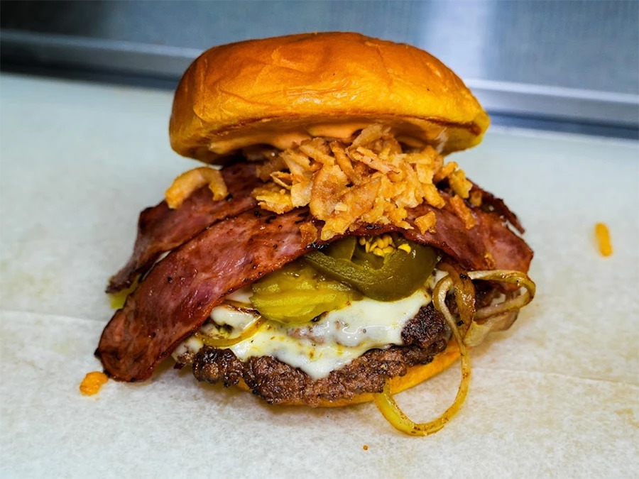 A mouth-watering gourmet burger with crispy bacon, melted cheese, jalapeños, and fried onions on a brioche bun, showcasing delicious offerings from Kingsbridge Estates Burger Restaurants.