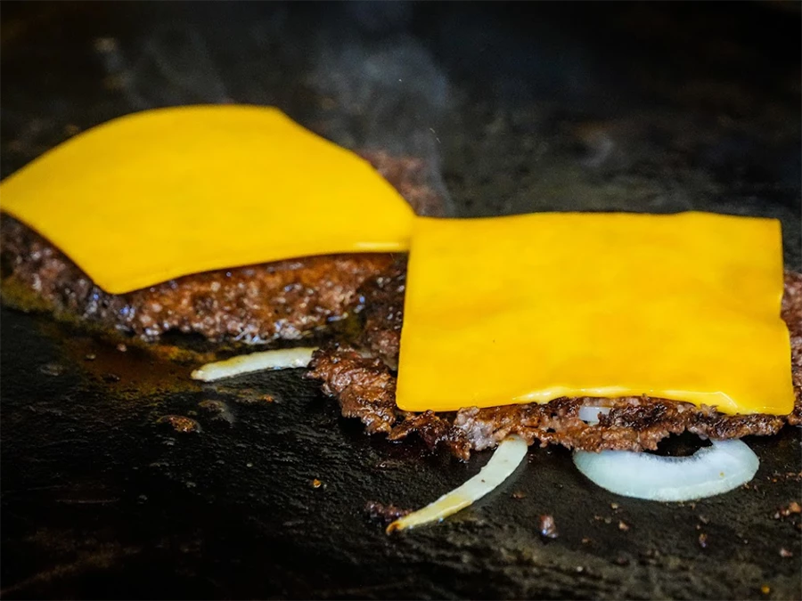 Two sizzling burger patties with melting cheese and onions on a griddle, representing the freshly cooked quality found at Top Burger Joints in Kingsbridge Estates