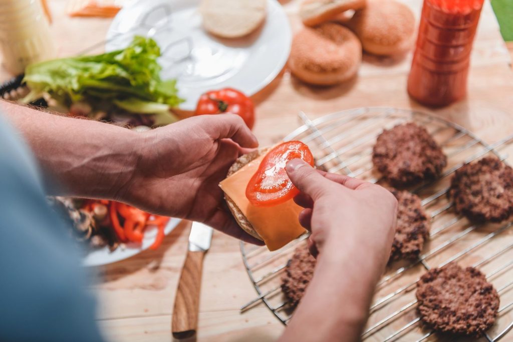 Hands assembling a best cheeseburger in Sugar Land with tomato, cheese, and fresh buns on a wooden table.