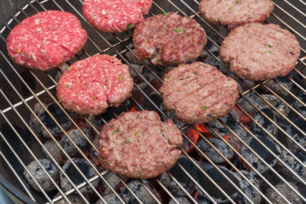 Raw and grilled best burgers in Sugar Land on a charcoal grill, featuring seasoned patties over glowing coals for an authentic backyard flavor.