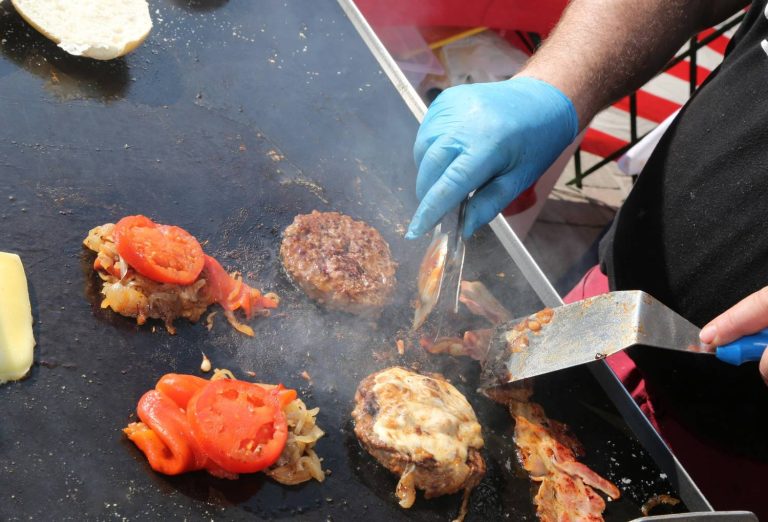 Chef using tongs and spatula to cook juicy burgers with onions, tomatoes, peppers, cheese, and bacon at burger restaurants Sugar Land, creating flavorful gourmet meals.