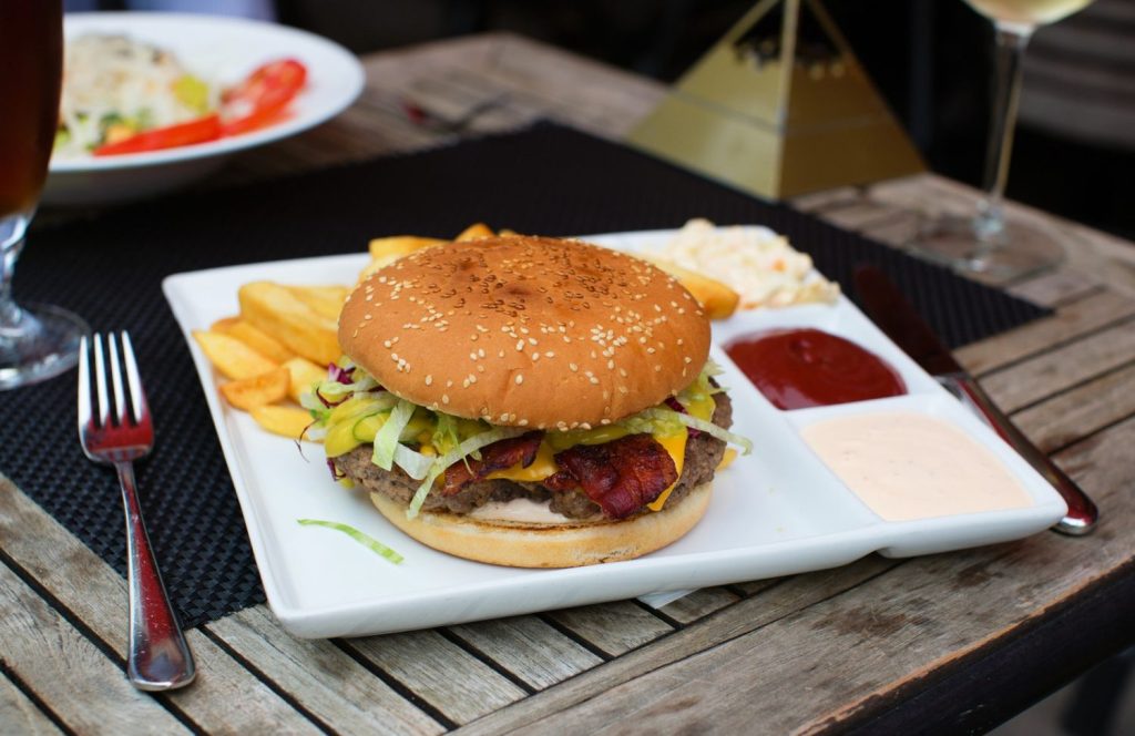 Plated affordable burger with bacon, cheese, lettuce, and pickles in Sugar Land, TX, served with fries, sauces, and coleslaw on wooden table.